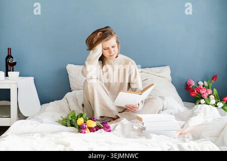 Happy smiling woman sitting on the bed wearing pajamas, with pleasure enjoying flowers and reading a book Stock Photo