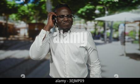 Man hand to ear touching glasses and ear in a white shirt on a busy street with cafe umbrellas and trees visible; confused. Stock Photo
