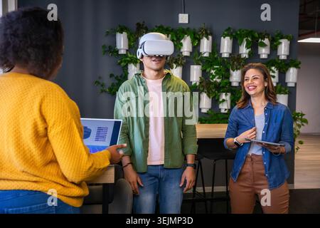 Diverse coworkers in mustard sweater testing VR headset and tablet, pointing to laptop at office. Collaboration, presentation, i Stock Photo