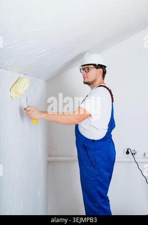 Young man in blue overalls and white hard hat painting wall into white color with a roller Stock Photo