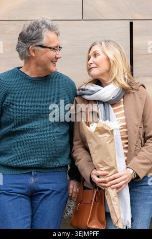 Senior couple in teal sweater and tan coat standing by stone wall, holding bouquet and handbag. Elderly, urban, denim, scarf, fl Stock Photo