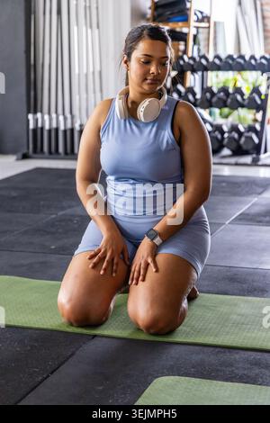 Asian woman kneeling and resting on green mat in gym, wearing light blue sportswear and headphones. Fitness, training, focus, du Stock Photo