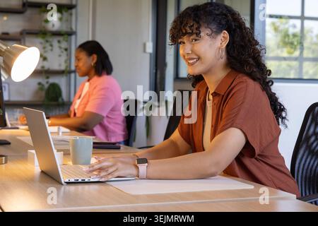 Diverse female coworkers smiling and working at long wooden desk with silver laptop, smartwatch. Coworking, office, openplan, na Stock Photo