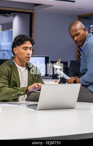 Diverse coworkers collaborating, reviewing printed charts and tablet using laptop at meeting table. Teamwork, graphs, reports, o Stock Photo