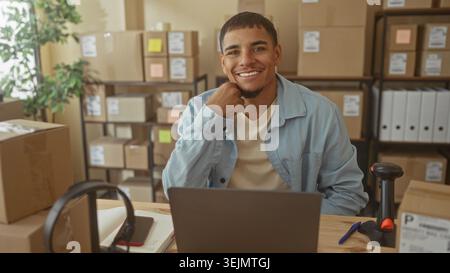 Man with hand on chin at desk with laptop and barcode scanner, surrounded by boxes and shelves in building; small business confidence. Stock Photo