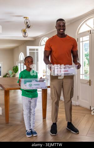 African American father and son holding clear recycling bins labeled GLASS and PAPER at home entry. Family, ecofriendly, sustain Stock Photo
