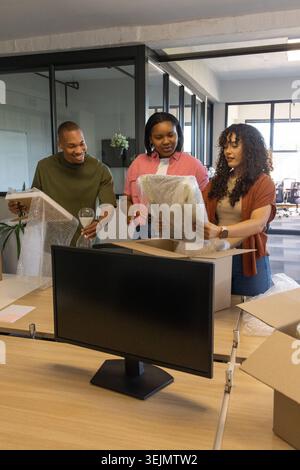 Diverse coworkers unpacking and inspecting wrapped decor and framed art by monitor at modern office. Teamwork, organization, pla Stock Photo