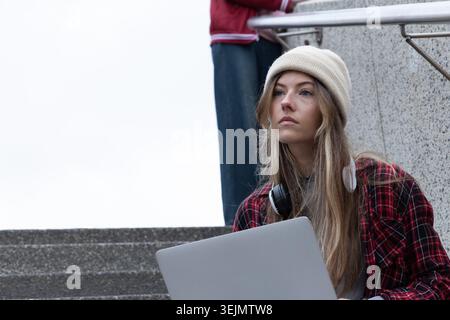 Woman and passerby sitting on urban stairway using laptop, plaid shirt and headphones, copy space. Notebook, earbuds, remote, ha Stock Photo