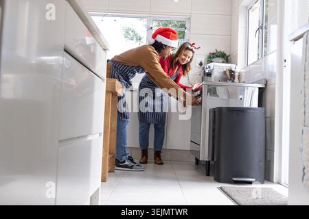 Couple opening stainless oven at home, wearing striped aprons, Santa hat and reindeer antlers. Kitchen, teamwork, festive, daylight, range, candid, gl Stock Photo
