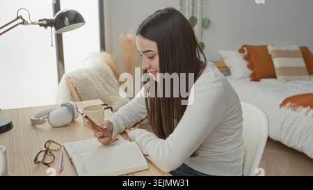 Young woman holding smartphone and studying with open notebook, pen, hand visible on phone at desk in studio with lamp and headphones; focused study. Stock Photo