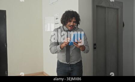Man holding blue tablet, fist pump while smiling in building hallway wearing shirt and tie checking property listing on screen; joyful success. Stock Photo