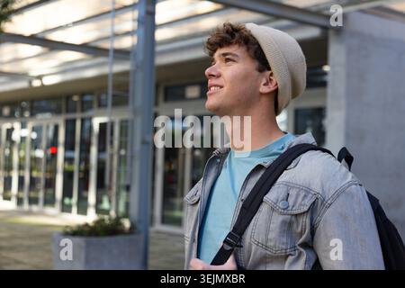 Teenage male standing at campus entrance wearing beige beanie and denim jacket, carrying backpack. Urban, university, student, g Stock Photo
