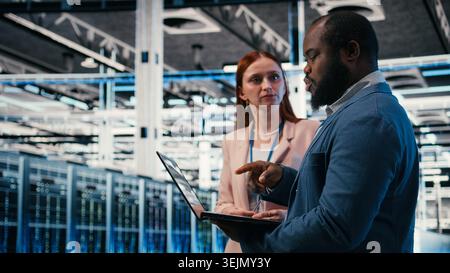 Engineering team solving tasks in server farm using laptop, doing network management virtualization. Teamworking data center colleagues oversee IT infrastructure supercomputers with notebook, camera A Stock Photo