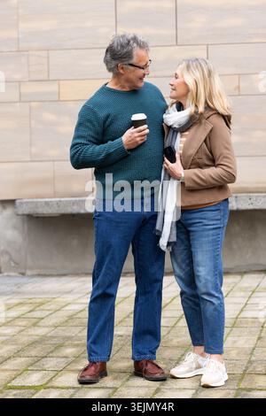 Senior couple standing close in urban plaza in jeans and sweaters holding coffee and sunglasses. Elderly, duo, city, bench, cup, Stock Photo
