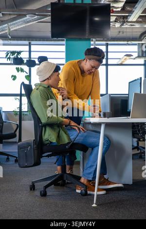 Asian man and African American woman collaborating at desk, leaning over, pointing at laptop. Collaboration, teamwork, office, modern, daylight, monit Stock Photo