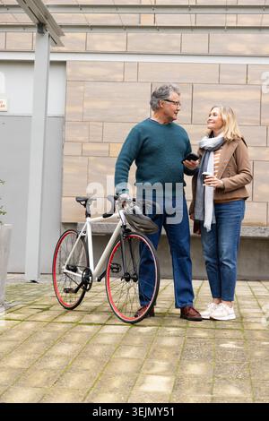 Senior couple standing in courtyard wearing casual jackets and scarf, holding bicycle and coffee. Plaza, bike, smartphone, knitw Stock Photo