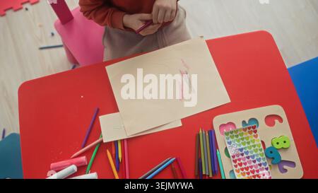 Boy tracing a hand and holding a red crayon while coloring on paper at a red kids table in a studio with stickers and colored pencils; concentration. Stock Photo