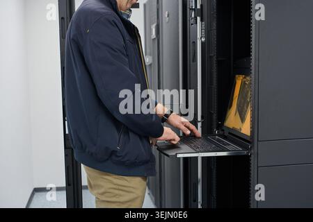 An IT technician types on a pull-out laptop console while servicing rack-mounted servers in a secure data center, depicting network maintenance, hardw Stock Photo