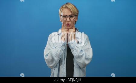 Caucasian blond woman in glasses pressing index fingers to her cheeks while puffing cheeks in a blue studio; playful mood. Stock Photo