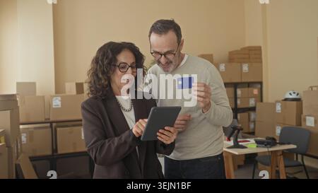 Man and woman pointing at tablet while man holds creditcard inside a storage building; efficient collaboration trust. Stock Photo