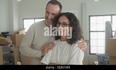 Man hugs woman with hand on shoulder amid packed moving boxes in building; contentment togetherness. Stock Photo