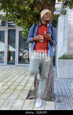African American man leaning against tree in plaza holding coffee and smartphone with headphones. Urban, casual, commuter, beanie, denim, joggers, bac Stock Photo