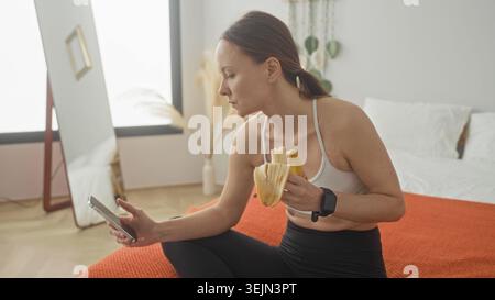 Young woman in sports bra holding a peeled banana and checking a smartphone while seated on bed in studio; post workout snack calm. Stock Photo