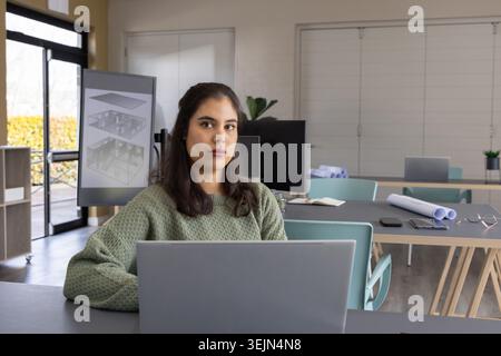 Woman sitting at office desk using laptop with tall monitor showing 3D plans and blueprints. Architect, designer, renderings, floorplan, schematic, mo Stock Photo