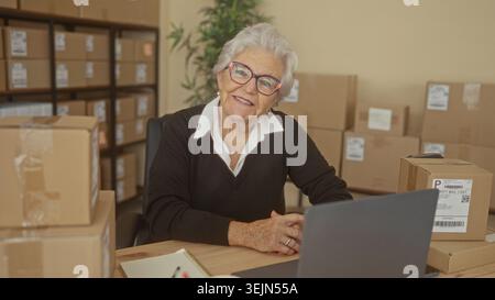 Elderly woman smiling with hands clasped by laptop and packing box in a building small business shipping station, surrounded by stacked parcel boxes; Stock Photo