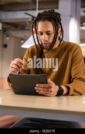 African American man sitting at wood table in brown quilted sweater, using tablet with stylus. Dreadlocks, coworking, cafe, modern, warm, lighting, pe Stock Photo