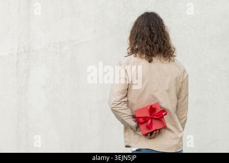Man hiding red gift box behind his back against white wall Stock Photo