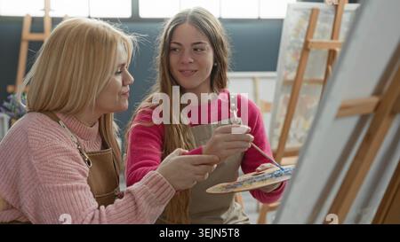 Woman painter and woman apprentice in aprons, blonde leaning in while the mentor points with brush to canvas and holds palette at easel in art studio; Stock Photo