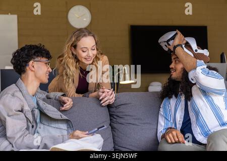 Diverse team sitting on gray sofa in office wearing blazer holding smartphone lifting VR headset. Coworking, collaboration, tech Stock Photo