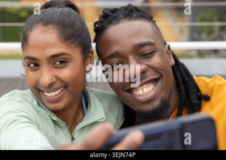 Diverse couple smiling and leaning while taking selfie with smartphone on bench in sportswear. Duo, lifestyle, candid, fitness, Stock Photo