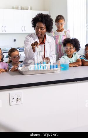 African American teacher labcoat pouring blue into tubes at school lab children watching in goggles. Educator, students, science Stock Photo