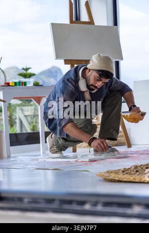 Man crouching on light floor in studio, holding wooden bowl, painting on floor canvas with easel. Artist, creativity, daylight, Stock Photo