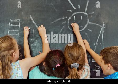 Four children use chalk to draw a smiling sun and simple shapes on a classroom chalkboard, collaborating in a playful, creative school activity that s Stock Photo