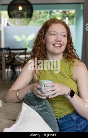 Red-haired woman sitting on light sofa holding pale green mug, wearing lime-green top and jeans. Cozy, home, livingroom, openpla Stock Photo