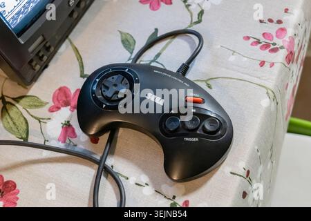 Black Sega Mega Drive control pad on floral tablecloth beside an old CRT television, wired retro game controller close-up for classic 1990s console ga Stock Photo