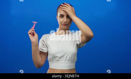 Woman with shaved short hair holds a pink razor while right hand rests on forehead in studio, wearing white crop shirt; contemplation identity. Stock Photo