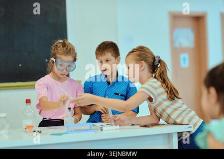 Three elementary students collaborate on a hands-on science experiment in a bright classroom, using goggles and simple lab equipment while practicing Stock Photo