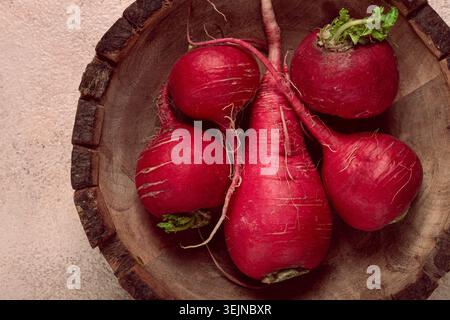 Freshly harvested red radishes displayed in a rustic wooden bowl on a textured surface Stock Photo