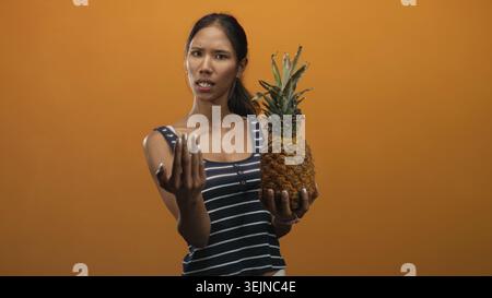 Thai woman brunette wearing navy striped tank holds a ripe pineapple in left hand and beckons with right palm forward on an orange studio set; skeptic Stock Photo