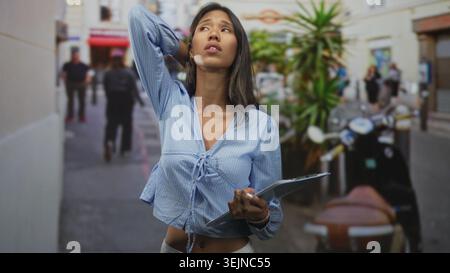 Thai woman holding clipboard with eyes closed touching neck on a narrow urban street; quiet serenity. Stock Photo