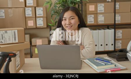 Woman smiling at laptop with folded arms inside a small business building, packages and shelves visible, barcode scanner on desk; small business confi Stock Photo