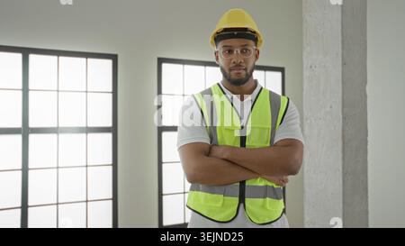 Man in yellow hardhat and neon safety vest gives thumbs up with one hand and arm crossed, smiling inside building under construction; confidence profe Stock Photo