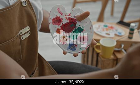 Man painter holds paint palette and brush with fingertip through palette hole, mixing paint while seated at an easel in studio; concentration. Stock Photo