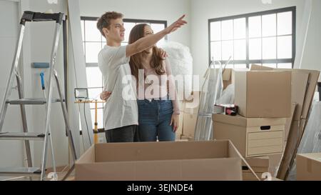 Woman and man hug among stacked cardboard boxes and ladder in an apartment while unpacking, woman gestures toward window and points; hopeful beginning Stock Photo