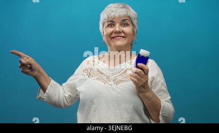 Senior woman with grey hair and plus size holding a blue medicine bottle points finger and smiles in studio; wellness confidence. Stock Photo