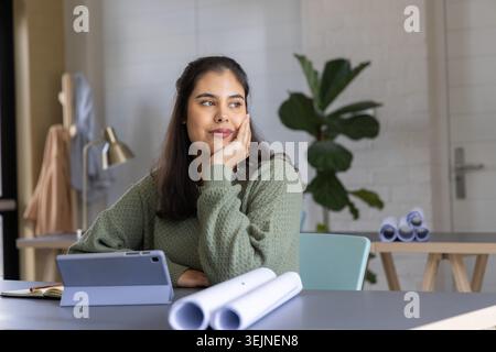 Asian woman in twenties wearing green sweater, reviewing tablet and rolled plans in home office. Studio, workspace, modern, mini Stock Photo